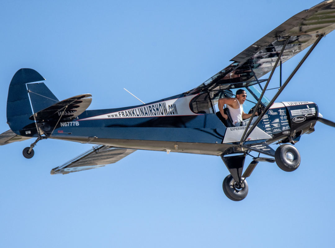 A Franklin’s Flying Circus biplane in flight with a wingwalker leaning out from the cockpit, wearing a white tank top and headband, against a clear blue sky; the aircraft displays “franklinairshow.com” along the fuselage.