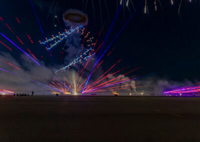 Nighttime air show performance featuring red and blue laser beams projecting across the airport ramp beneath fireworks and illuminated aerial effects. Smoke drifts across the performance area while spectators watch from a distance, creating a dramatic finale against the dark evening sky.