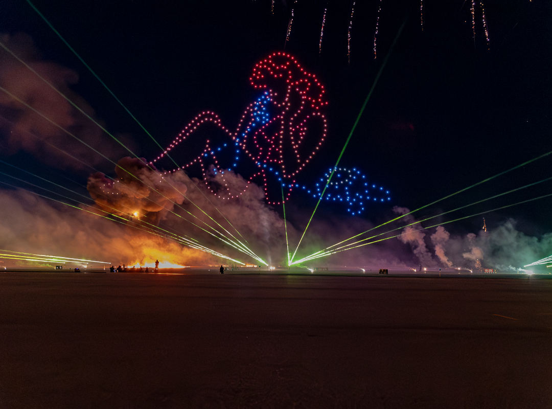 Laser Encore nighttime performance at Wings Over Batavia Air Show, featuring a large drone light display of a reclining pin-up figure outlined in red, white, and blue lights above the airfield. Green laser beams radiate across the tarmac while pyrotechnics, smoke effects, and fireworks illuminate the night sky.