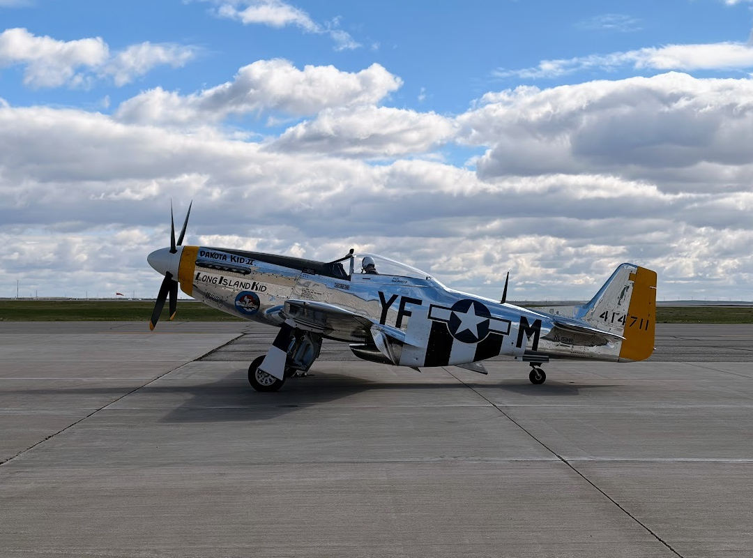 P-51 Mustang Dakota Kid II parked on an airport ramp beneath a cloudy sky. The polished silver aircraft displays WWII-era U.S. Army Air Forces markings, with both "Dakota Kid II" and "Long Island Kid" painted on the nose and a yellow tail section visible in profile.
