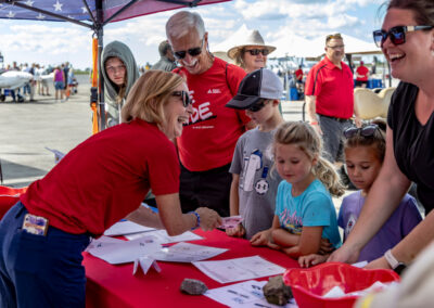 Children and adults gather around a Discovery Zone activity table as a volunteer hands out materials beneath a tent at the Wings Over Batavia Air Show.