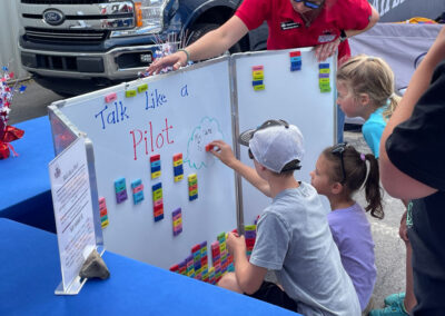 Children place colorful aviation words on a magnetic “Talk Like a Pilot” board while a volunteer assists nearby.