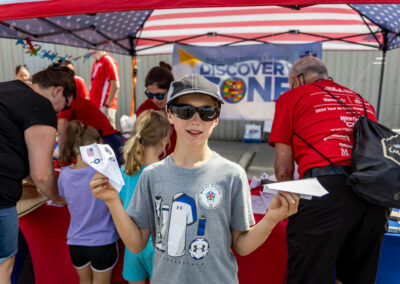A child wearing sunglasses holds up two completed paper airplanes in front of the Discovery Zone tent.
